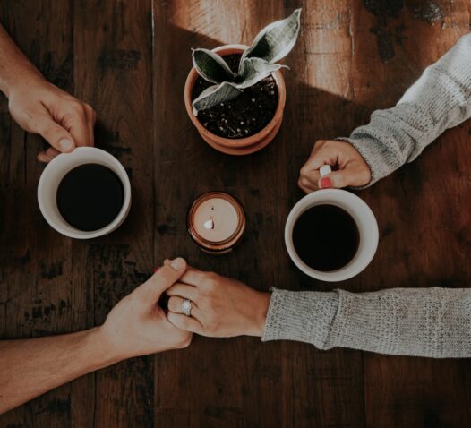 Två personer håller varandra i händerna vid ett bord med kaffe och en växt.
