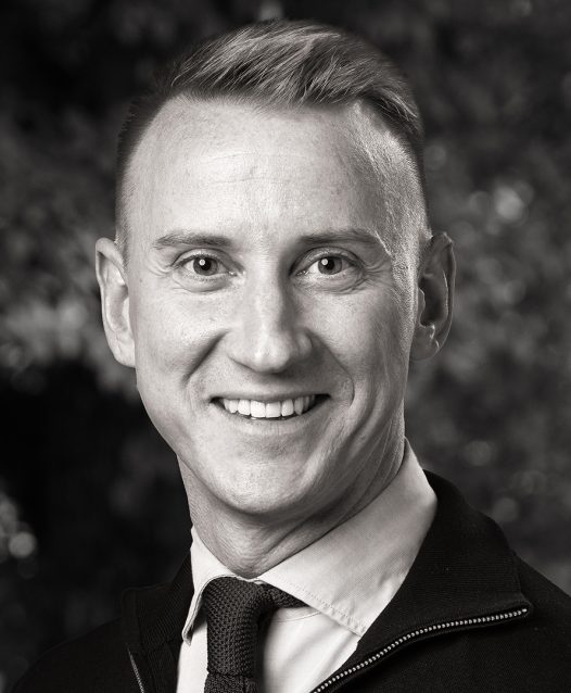 Man som ler, iklädd skjorta och slips, med suddig bakgrund av träd.